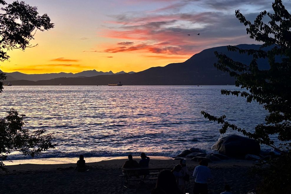 A colourful Vancouver sunset at Kits Beach, with silhouettes of people on the sand, tankers on the water, and the North Shore mountains against the sky. (Bhagyashree Chatterjee/Daily Hive)